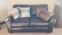 Full view of dark brown faux leather loveseat with four throw pillows (two plaid, two mosaic).