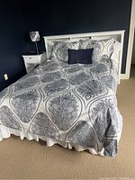 Full made bed showing white bed frame with headboard, patterned bedding, decorative pillows, and dust ruffle on box spring