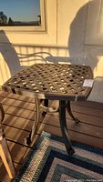 Metal outdoor side table with lattice pattern top, bronze finish, on deck with sunlight and shadow.