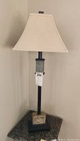 Full view of one lamp showing white square fabric shade, black metal rod, and stone base on black square platform.