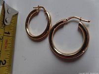 Two gold hoop earrings next to measuring tape showing approximate 1″ size