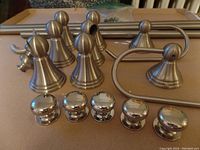Overview of bathroom hardware pieces laid out, showing mounting bases with round finials, chrome cabinet knobs, toilet paper holder hooks, and metal rods.