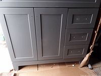 Grey bathroom vanity cabinet showing front with closed doors and three closed drawers on the right side. Minor damage visible on right side panel.