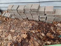 Stack of about 30 rectangular concrete interlocking pavers outdoors against a house wall, surrounded by dry leaves.