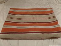 Folded cotton area rug showing orange, beige, brown and blue stripes.