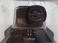 Close-up of the black rectangular action camera lens inside a clear waterproof protective case, with a sticker on the lens saying 'Please remove me before using'.