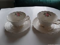 Two white porcelain tea cups with pink rose floral designs on matching saucers, displayed on a white surface.