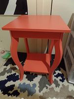 Coral side table with rectangular top, curved legs and lower shelf, placed on a multicolored rug.