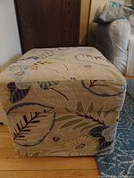 Closed rectangular storage ottoman covered with removable floral fabric with leaf and flower pattern in muted blues, greens, and browns.