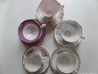 Top view of five tea cups and saucers displaying their floral designs and varied colors including pink, purple, white, and grey with gold accents
