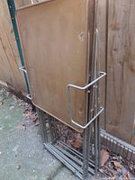 Four stacked metal folding tables with signed trays leaning against a fence outdoors