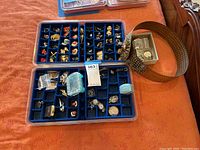 Two blue plastic compartment trays filled with assorted jewelry items alongside a perforated metal waist belt.