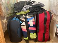 Pile of four travel bags including multicolor hard-shell spinner, red soft duffel, navy blue soft bag and partially visible clothing