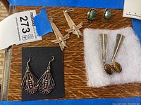 Photo showing six pairs of earrings arranged on wooden surface with part of blue and white MaxSold tags visible.