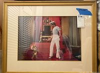 Full front view of framed cel showing gold-finish frame, off-white mat, and central image.