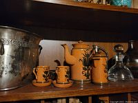 Shelf with the orange ceramic tea set, including teapot, smaller pot, two cups and saucers. Beside them is a large metal decorative jar with embossed design and two clear glass decanters.