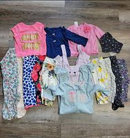 Photo showing an assortment of baby and toddler girl clothes including tops, leggings, and a hoodie, arranged on wooden floor.