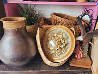 Photo showing large metal vase with wicker top trim, wicker baskets behind, round wooden flower frame and bird statue