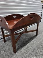 Side angled view of the wooden butler's tray table showing the curved sides and cut-out hand grips of the removable tray and the crossed leg support structure.
