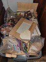 Wide view of container filled with various woodcraft items including wooden numbers, heart-shaped box, wooden frame, sticks, and bags of other wooden craft components.