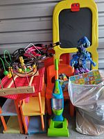 Photo showing a variety of children's toys including a colorful plastic playhouse, a wooden bead maze, a toy vacuum cleaner, a yellow-framed chalkboard, and a blue dinosaur figure
