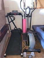 Treadmill, elliptical bike, pull-up bar, dumbbells, weight plates and foam roller arranged by a wall and sofa