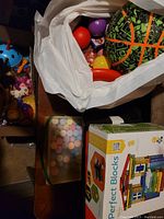 Photo showing a white plastic bag holding various colorful balls and an inflatable basketball, a box of Perfect Blocks, container of sidewalk chalk, and other small toys around.