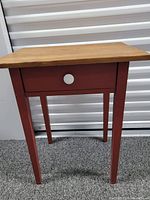 Front view of wooden side table with red legs and drawer front, natural wood top and white round knob on drawer.