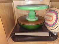 Green wooden cake stand atop large metal bowl, brown tray below, and woven mat beside them inside a wooden cabinet