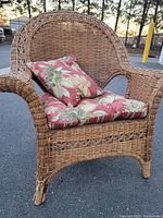 Front side view of brown wicker chair with floral cushions. Shows intact wicker with decorative weaving around seat. Right front leg is damaged at bottom corner.