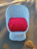 White wicker chair with a red cushion pillow outdoors on pavement under sunlight, viewed from front and slightly top angles.