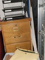 Wooden two-drawer filing cabinet with brass handles, showing light wood grain and label frames. Black metal mesh trays and some file folders on top and next to the cabinet.