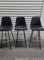 Set of three high metal bar stools with black upholstered seats and backs, arranged in a row against a roller door.