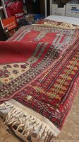 Image showing one red and cream wool area rug draped partially, displaying intricate geometric patterns, fringe at the end, and some surface wear.