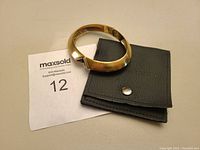 Gold bracelet placed on a black branded pouch with snap closure and a MaxSold lot number card.