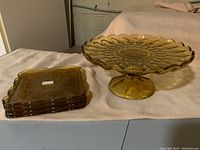 Photo showing stacked four square amber glass plates next to the amber footed cake stand on a cloth surface