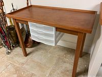 Front angle view of rectangular wooden table with raised edge tabletop and three white plastic drawers underneath.