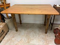 Rectangular solid wood drop leaf table with natural finish, turned oak legs, positioned against a white wall on tile flooring.