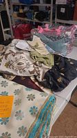 Large assortment of various fabrics including floral, black with gold motifs, and green fabric pieces on table with plastic bag holding more fabric
