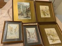 Five prints in frames, two larger with gold mats and three smaller with darker mats on table, showing street and architectural scenes.
