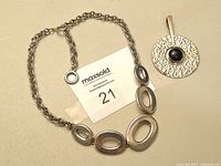 Photo of sterling silver necklace laid flat with oval-shaped hollow center links and round textured pendant with black stone beside it.