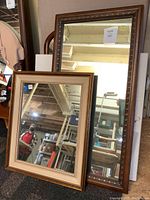 Two wooden framed mirrors leaned against a wall in a storage setting, one with a two-tone beige and brown frame, the other taller with decorative beading on dark brown frame.