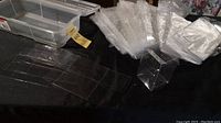 Box containing multiple clear, folded retail plastic packaging boxes on a dark surface next to a beige plastic storage container.