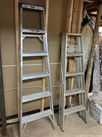 Two aluminum step ladders side by side, one taller (6 ft) and one shorter (5 ft) with fold-down paint tray visible on shorter ladder.