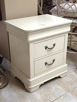 Front angled view of the painted wooden side table with two drawers, decorative metal handles, and slight marks on the surface.