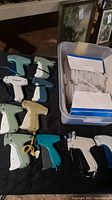 Nine retail tagging applicator guns in assorted colors arranged beside a box of white plastic tags on a black surface.