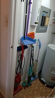 View showing broom with red grip handle, Swiffer dry mop, blue extended dustpan and three stick vacuum cleaners arranged vertically against the wall near a utility area.