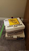 Photo of yarn cone winder SHW-10 box with yellow illustration and wooden spool holders behind