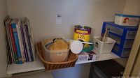 Shelf with craft books, assorted crafting supplies including wood forms, a basket with yarn, a plastic loom knitting kit, and cylindrical containers.