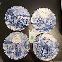 Four Royal Delft blue and white hand-painted plates arranged on a black surface showing various Dutch countryside and village scenes, measuring about 8 inches each.